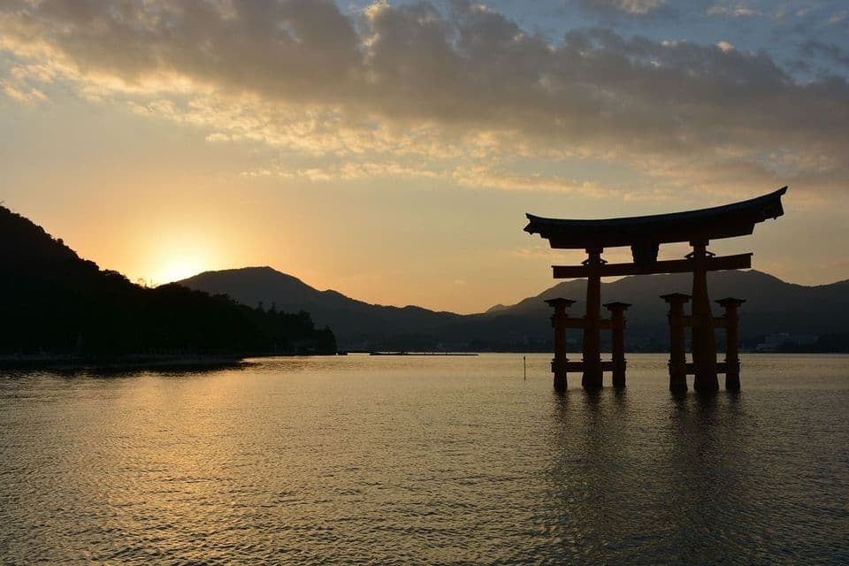 Un tradizionale torii giapponese si staglia sull'acqua al tramonto, con il sole che cala dietro montagne lontane sotto un cielo nuvoloso.