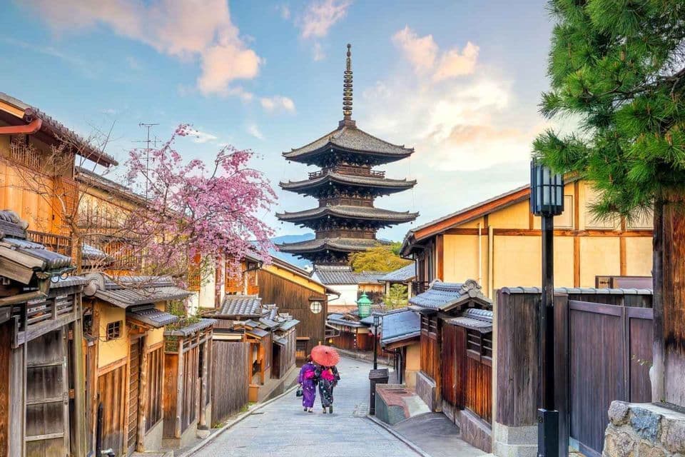 Due persone in kimono tradizionali passeggiano lungo una storica strada giapponese con un'alta pagoda e un ciliegio in fiore sullo sfondo.