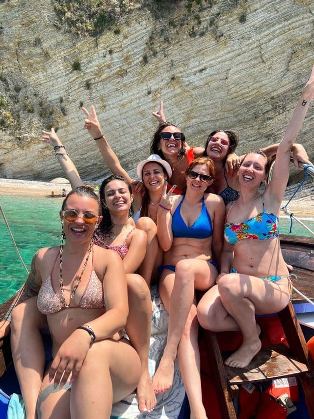 Eine WeRoad-Gruppenreise mit lächelnden Frauen in Bikinis, die auf einem Boot neben einer Felsklippe und türkisfarbenem Wasser posieren.