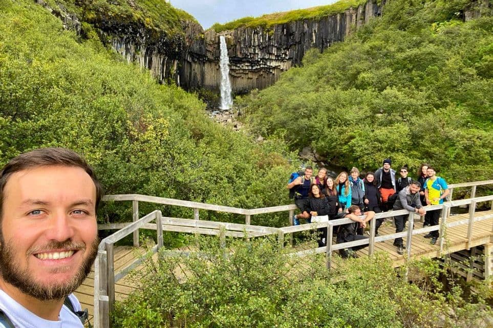 A man takes a smiling selfie with a WeRoad group trip on a wooden bridge, with a waterfall and basalt columns in the background.