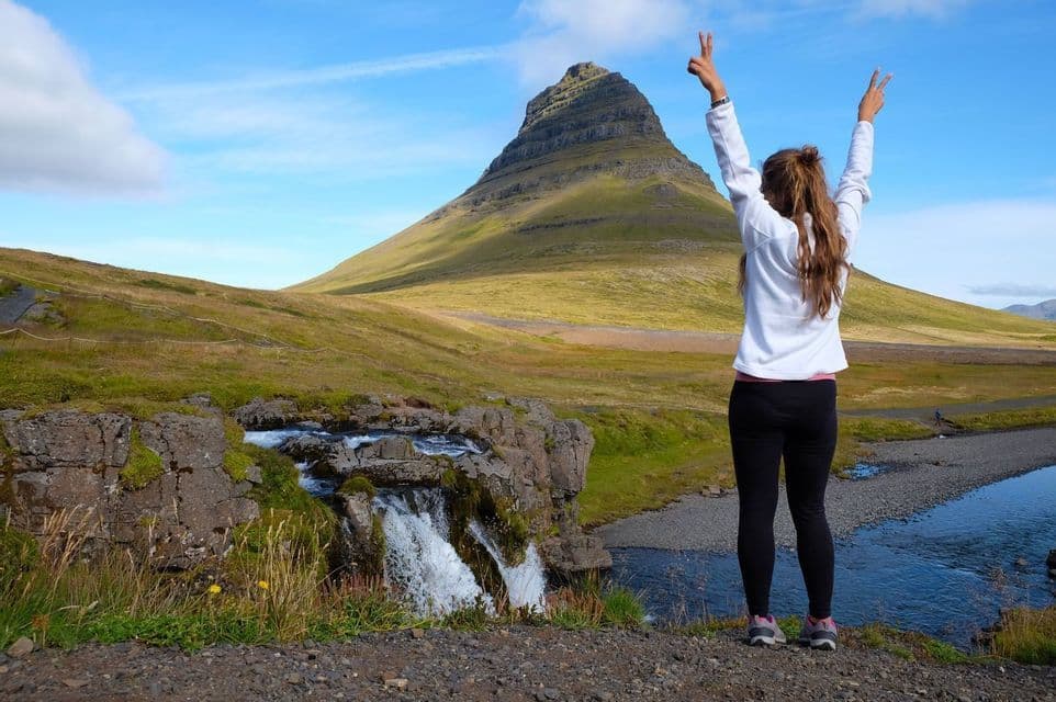 A woman stands with arms raised in victory signs, facing a green conical mountain and a small waterfall.