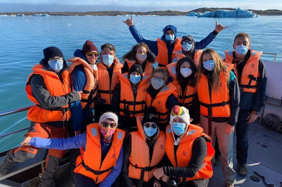 A WeRoad group trip wearing life vests and face masks poses for a photo on a boat, with icebergs in the background water.