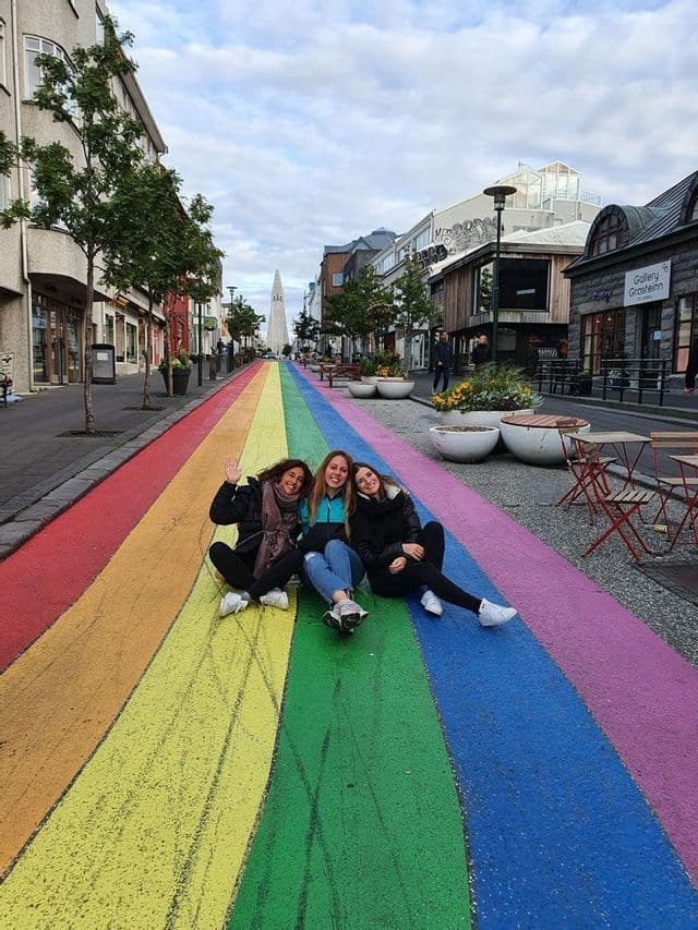 A WeRoad group trip of three women smiling while sitting on a long, straight road painted like a rainbow in a city.