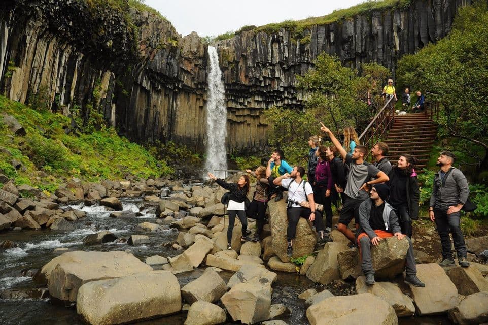 A WeRoad group trip sits on rocks by a stream, pointing up at a waterfall flowing over basalt columns.