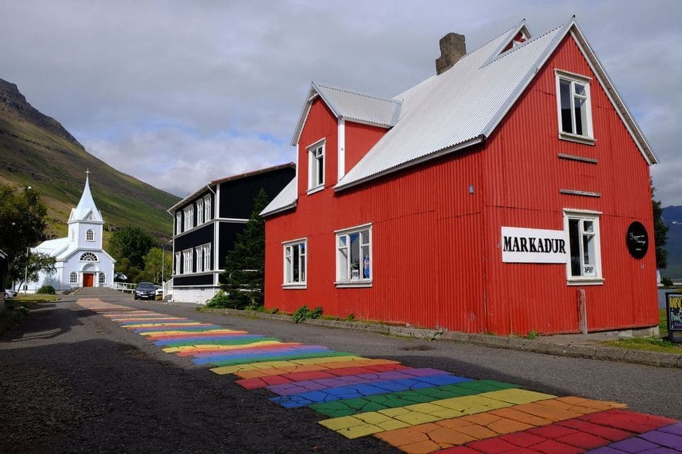 A street with a painted rainbow path leads past colorful buildings toward a white church at the base of a mountain.