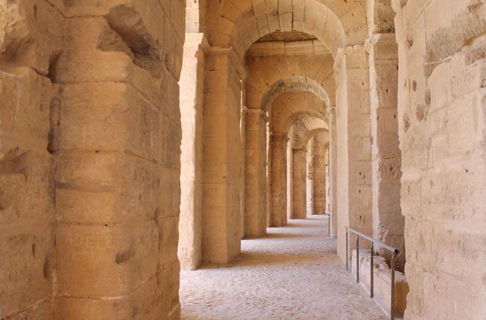 Un lungo corridoio vuoto all'interno di un antico anfiteatro in pietra, con una serie ripetuta di archi e pilastri che creano una prospettiva sfuggente.
