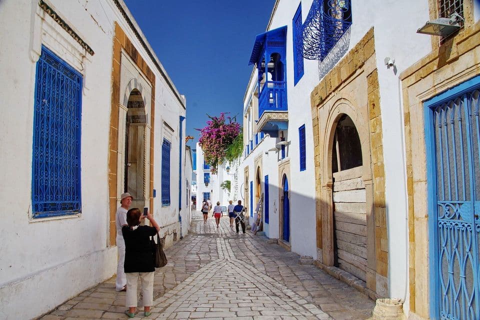 Le persone camminano lungo una stretta strada di ciottoli tra edifici imbiancati a calce con porte, finestre e balconi di un blu brillante.
