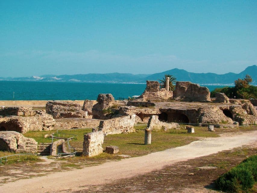 Antiche rovine in pietra su una costa erbosa che si affaccia su un mare blu calmo con montagne in lontananza.