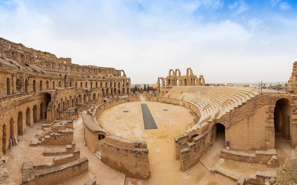 Una vista ampia delle rovine di un antico anfiteatro in pietra con gradinate a più livelli e un'arena centrale sabbiosa.