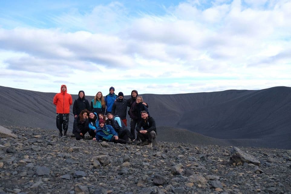 A WeRoad group trip posing for a photo on the rocky rim of a large, dark volcanic crater under a cloudy blue sky.