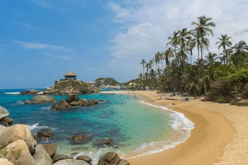 A scenic tropical bay with people swimming in turquoise water, a sandy beach lined with palm trees, and a hut on a rocky outcrop.