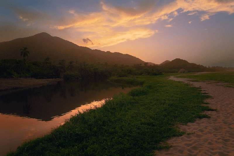 A river flows alongside a grassy bank and sandy beach, with mountains in the background under a sunset sky.