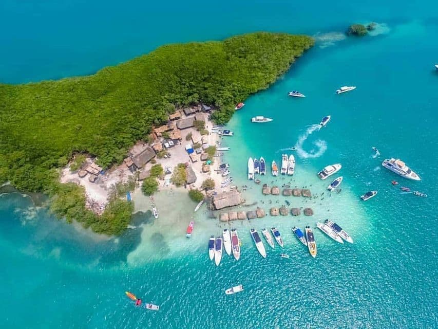 An aerial view of a small, lush green island with numerous boats anchored and people swimming in the turquoise water.