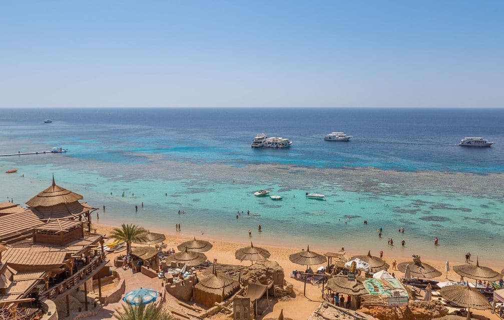 Una vista elevada de una playa tropical con cabañas de techo de paja, gente nadando en agua turquesa y barcos en el mar azul profundo.