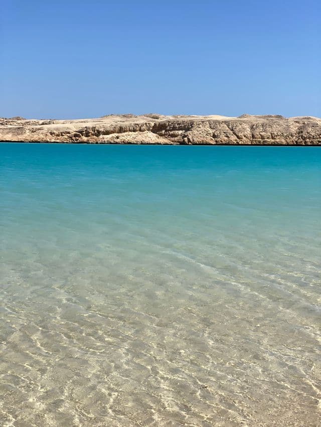 Agua turquesa cristalina ondula sobre un fondo arenoso, con una costa desértica rocosa al fondo bajo un cielo azul claro.