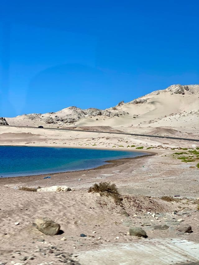 Una carretera costera bordea una bahía con agua turquesa y colinas desérticas de arena bajo un cielo azul claro.