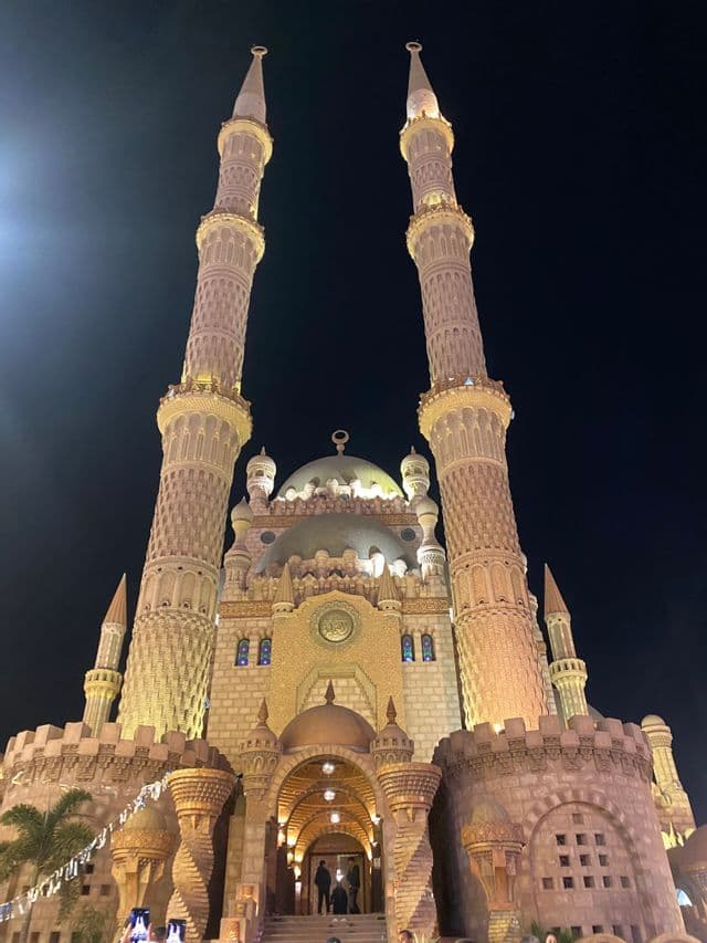 Una toma en ángulo bajo de una mezquita grande y ornamentada con dos minaretes y una cúpula central, brillantemente iluminada contra el cielo nocturno.