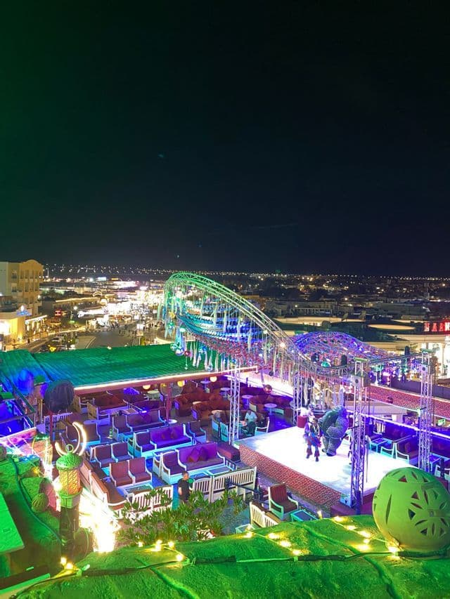 Una vista elevada de un vibrante bar en la azotea por la noche, con artistas disfrazados en el escenario y una ciudad iluminada de fondo.