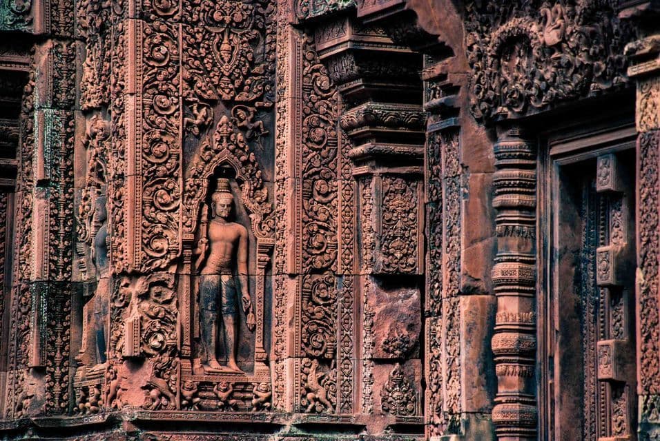 Close-up of intricate carvings on a reddish stone temple wall, featuring a standing deity in a decorative niche.