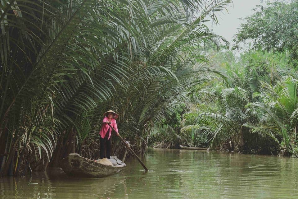 Une personne coiffée d'un chapeau conique et vêtue d'une chemise rose pagaie une petite barque en bois sur une rivière bordée d'une végétation luxuriante de jungle.