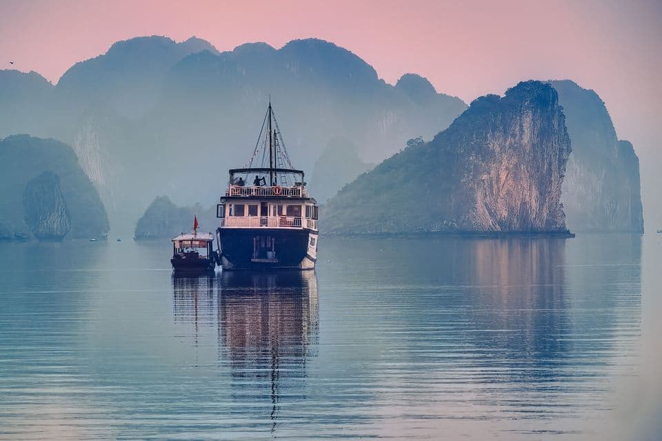 A WeRoad group trip on a tour boat floating on calm water among misty limestone karsts during a pink dusk.