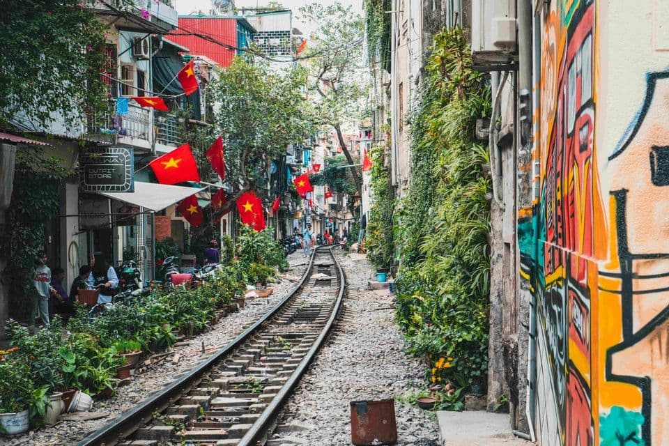 A railway track runs through a narrow urban alley lined with buildings, potted plants, red flags, and a colorful graffiti mural.