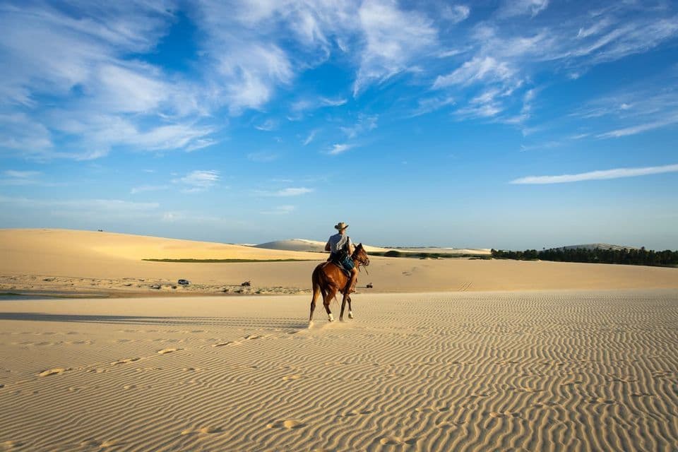 Una persona con un cappello cavalca un cavallo marrone attraverso vaste dune di sabbia ondulate sotto un cielo azzurro brillante con nuvole sottili.