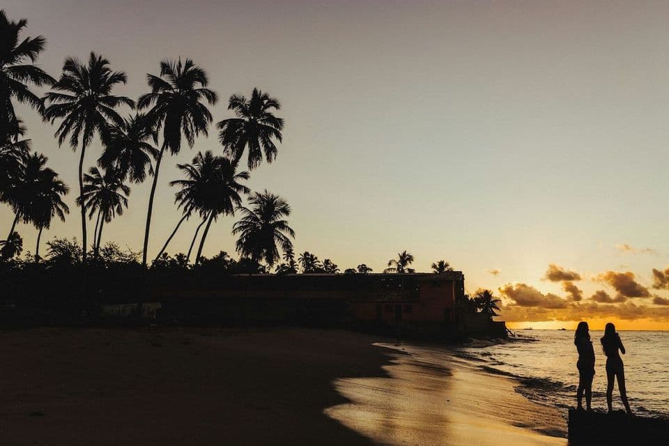 Sagome di due persone di un viaggio di gruppo WeRoad in piedi su una spiaggia al tramonto, con palme e l'oceano.