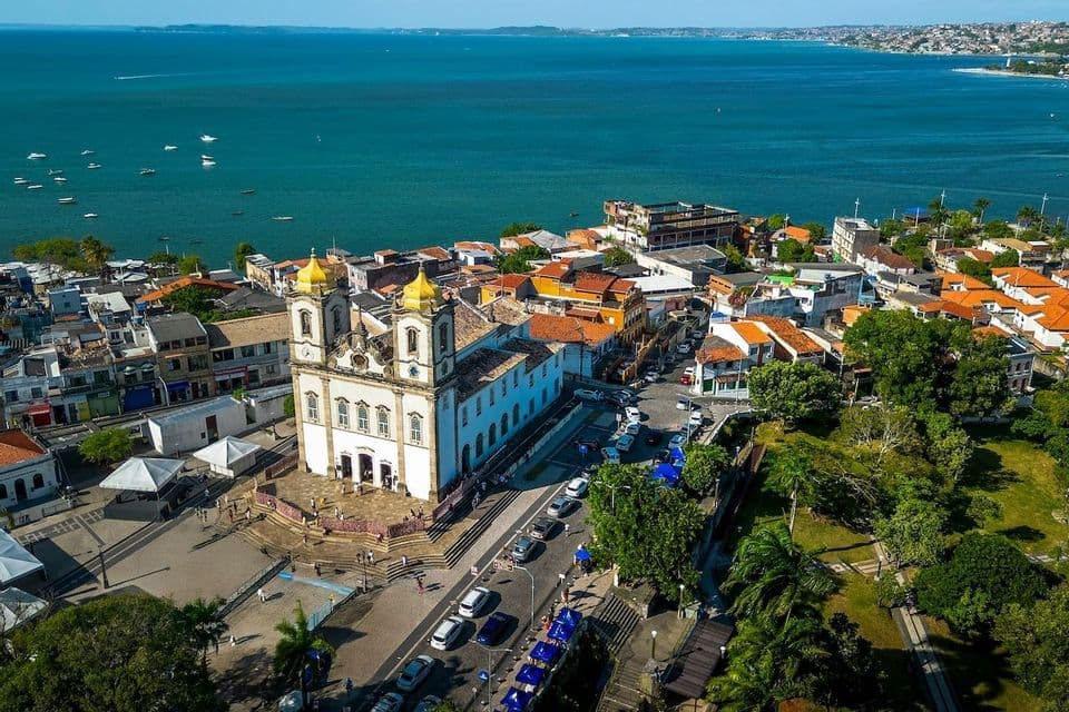 Una vista aerea di una chiesa bianca con due torri a cupola dorata in una città costiera che si affaccia su una vasta baia blu con barche.