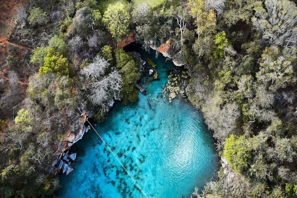 Una vista aerea dall'alto di una piscina naturale con acqua cristallina e turchese, circondata da una fitta foresta e sovrastata da una zipline.