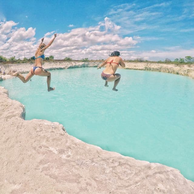 Two women in bikinis jumping from a rocky ledge into a pool of turquoise water under a blue, cloudy sky.