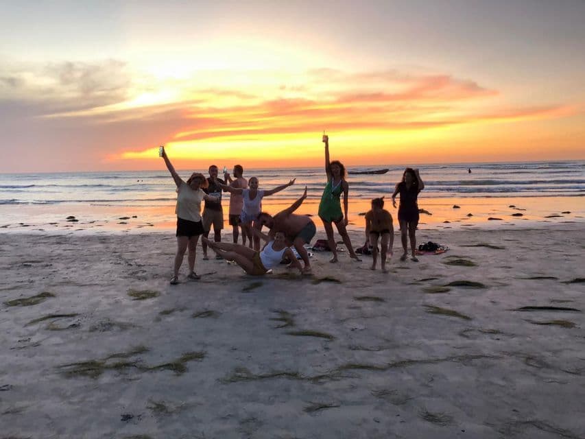 A WeRoad group trip of people posing playfully on a sandy beach with the ocean behind them during a colorful sunset.