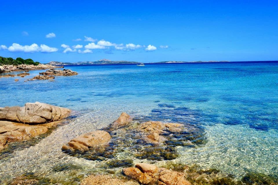 Una costa rocciosa incontra acque calme, limpide e turchesi sotto un cielo azzurro brillante con una singola barca in lontananza.