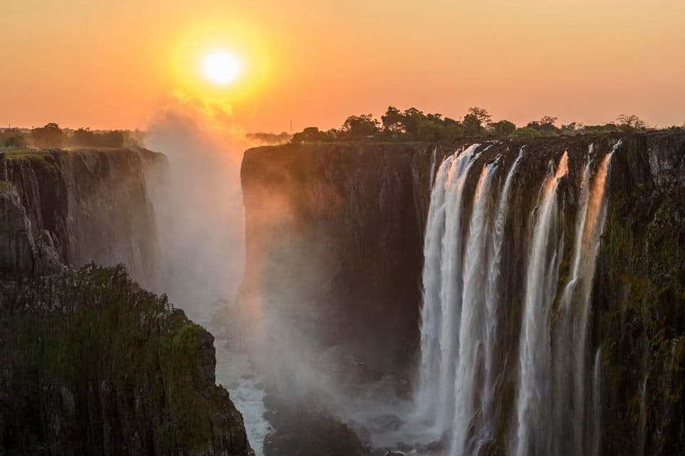 Una cascata imponente scende da una parete rocciosa in una gola profonda al tramonto, con la nebbia che sale dall'acqua sottostante.