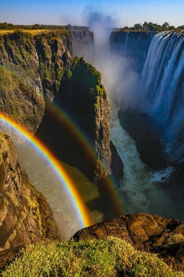 Un vivace doppio arcobaleno si inarca su un canyon profondo con una potente cascata che precipita nel fiume sottostante.