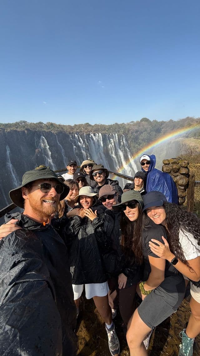 Un gruppo WeRoad si scatta un selfie sorridente in impermeabile davanti a una grande cascata con un arcobaleno nel cielo.
