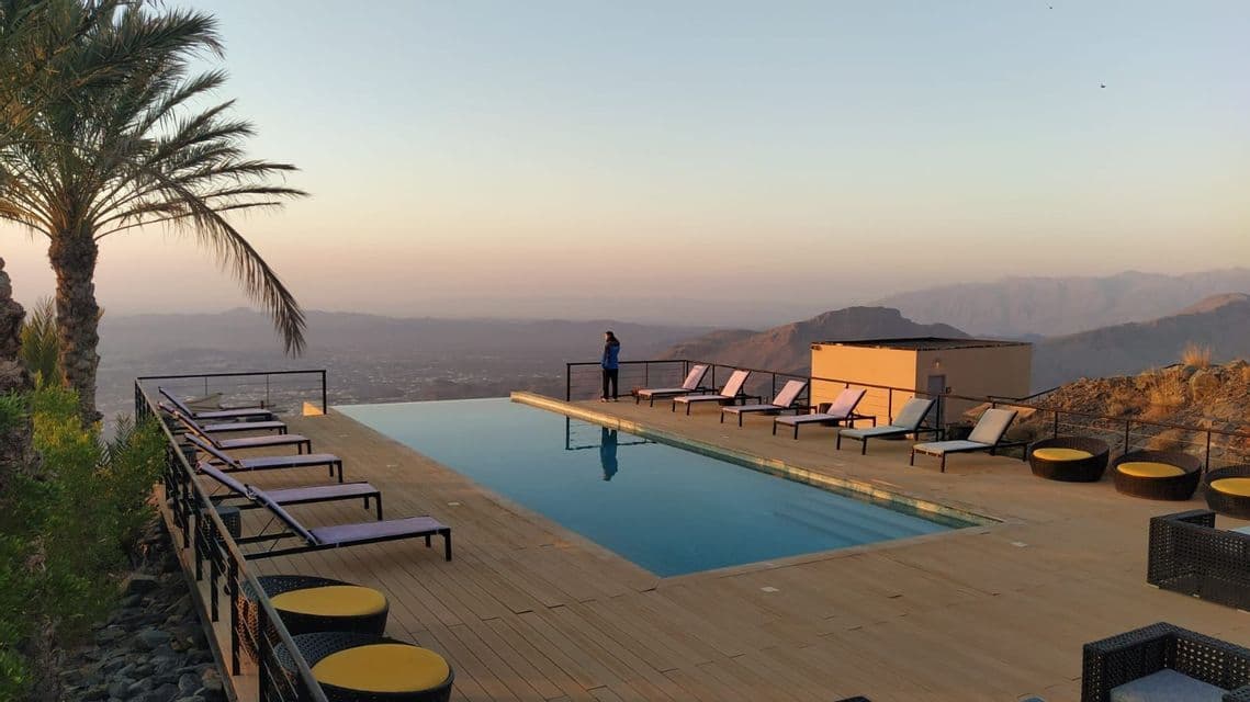 Una persona de pie junto a una piscina infinita en una terraza de madera, contempla un vasto paisaje montañoso al atardecer.
