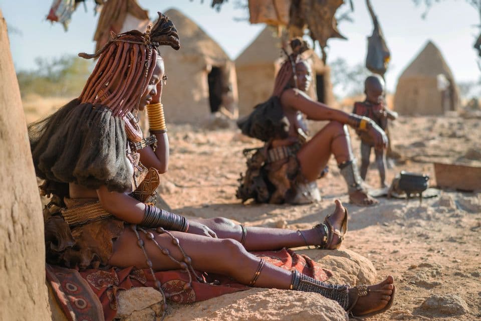 A woman in traditional attire with an intricate hairstyle sits on a rock outside a hut in a village.