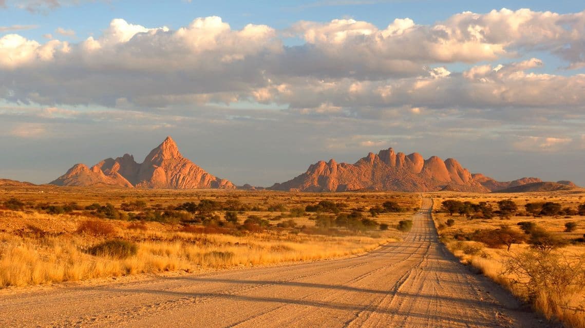 A long dirt road stretches through a golden savanna landscape towards rocky mountains under a partly cloudy sky.