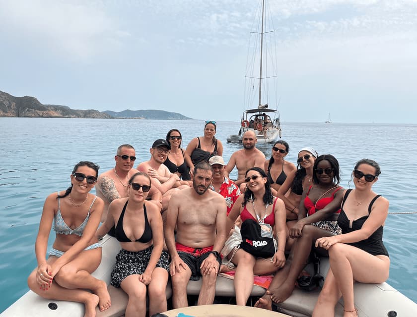 Un groupe WeRoad prend la pose pour une photo à la proue d'un bateau, avec la mer et un voilier en arrière-plan.