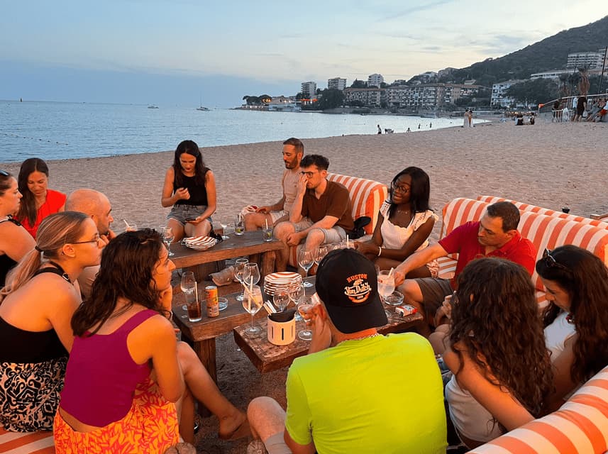 Un voyage en groupe WeRoad, les participants se détendent sur des canapés rayés au bord d'une plage, partageant des boissons autour de tables en bois au coucher du soleil.