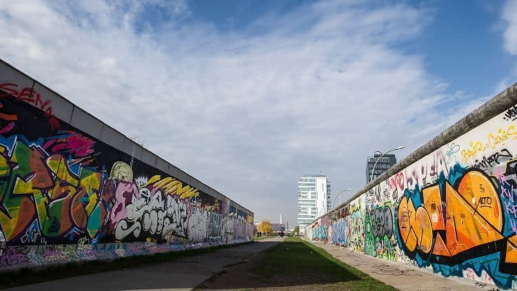 Due lunghe pareti di cemento ricoperte di graffiti colorati formano una passerella con edifici moderni visibili in lontananza sotto un cielo nuvoloso.
