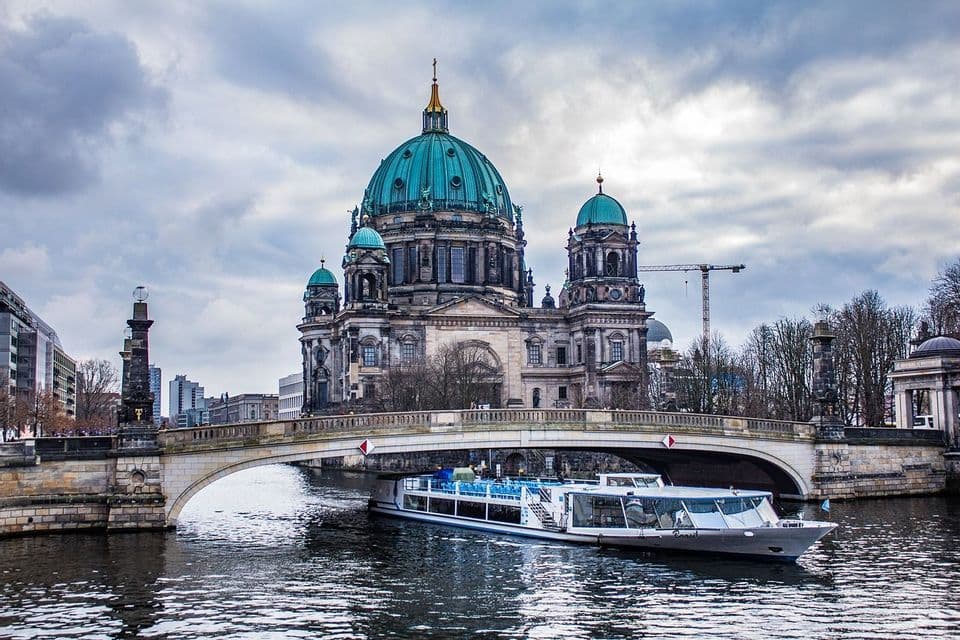 Una barca turistica naviga sotto un ponte di pietra su un fiume, con una grande cattedrale dalla cupola verde sullo sfondo.