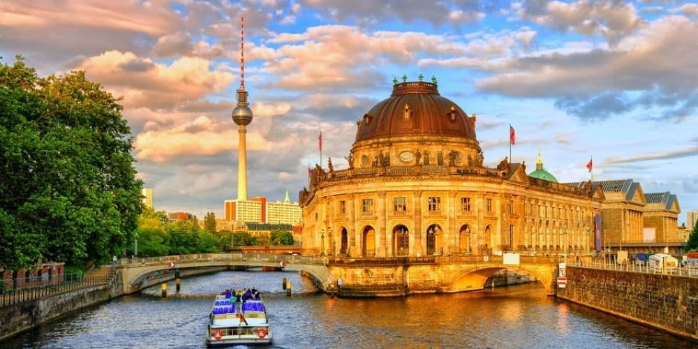 Una barca turistica naviga su un fiume cittadino, passando davanti a un palazzo dalla cupola ornata, con un'alta torre della televisione in lontananza al tramonto.