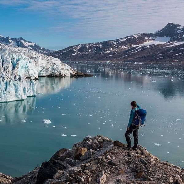 Un escursionista con uno zaino si erge su uno sperone roccioso che domina una baia con un grande ghiacciaio e iceberg galleggianti.