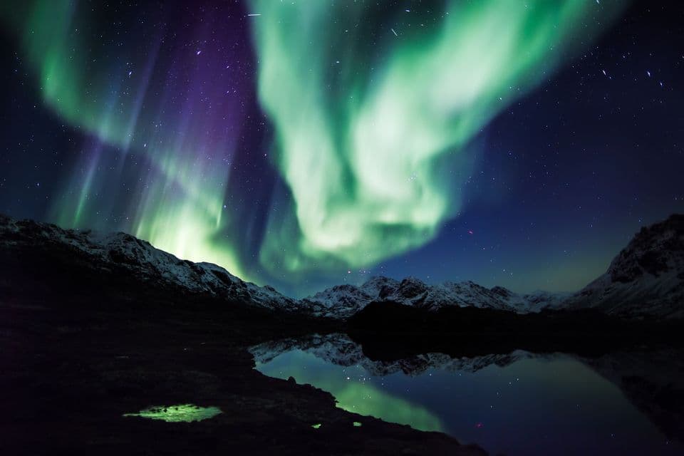 L'aurora boreale risplende di verde e viola sopra montagne innevate, il suo riflesso visibile in un lago immobile di notte.