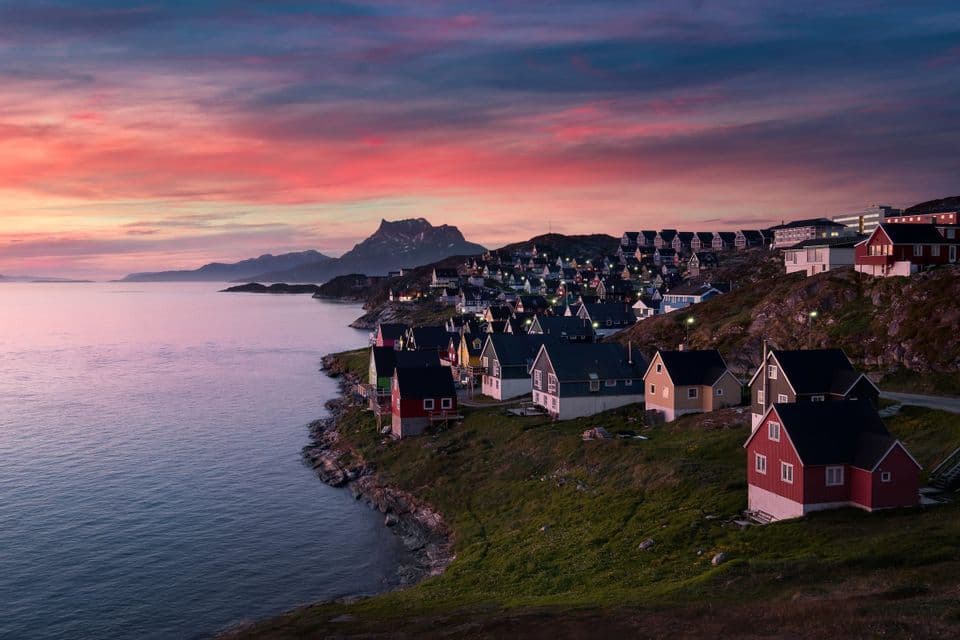 Un borgo marinaro con case colorate si adagia su una costa verde e rocciosa a ridosso del mare, sotto un vibrante cielo al tramonto.