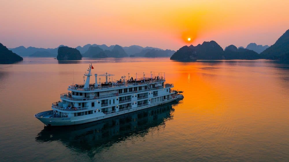 A multi-deck cruise boat carrying a WeRoad group trip sails on calm water between karst islands at sunset.