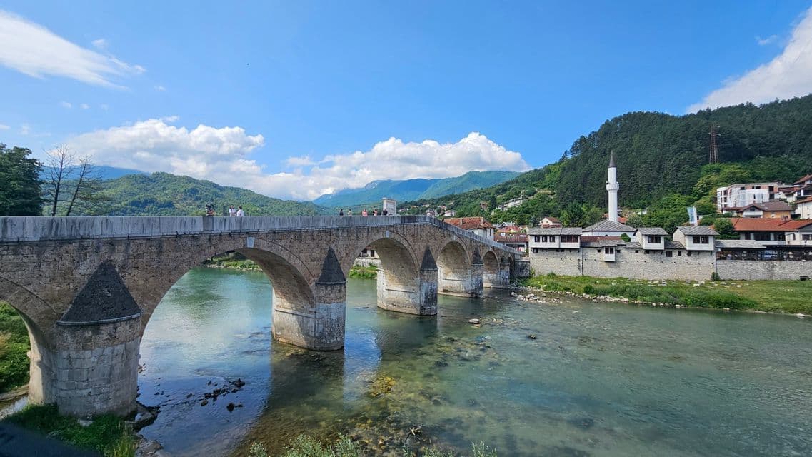 Eine große, mehrbögige Steinbrücke überspannt einen klaren Fluss neben einer Stadt mit einem Minarett und bewaldeten Bergen.