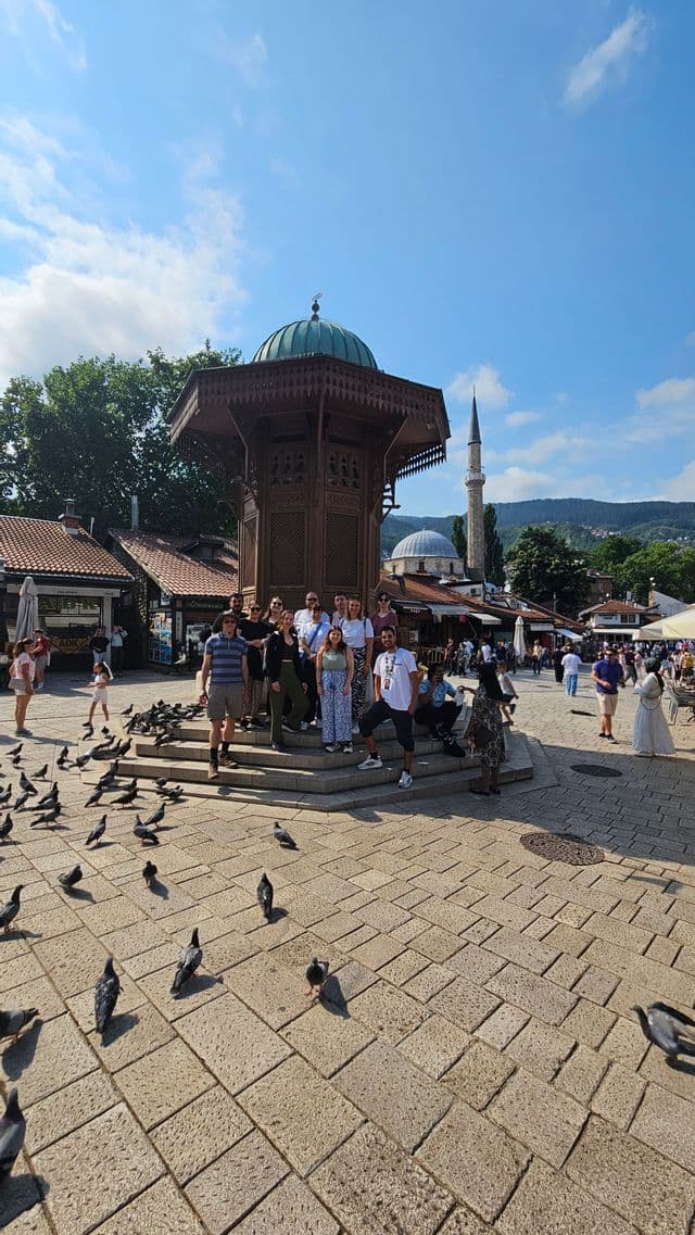 Eine WeRoad-Gruppe posiert an einem sonnigen Tag auf einem gepflasterten Platz mit einem verzierten Holzbrunnen und vielen Tauben.